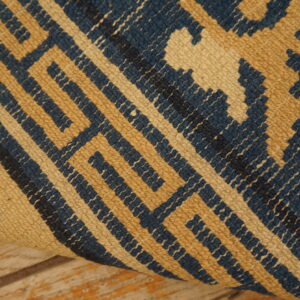 A closeup shows a rolled rug fragment featuring a geometric blue and tan repeating pattern over distressed wood floorboards.