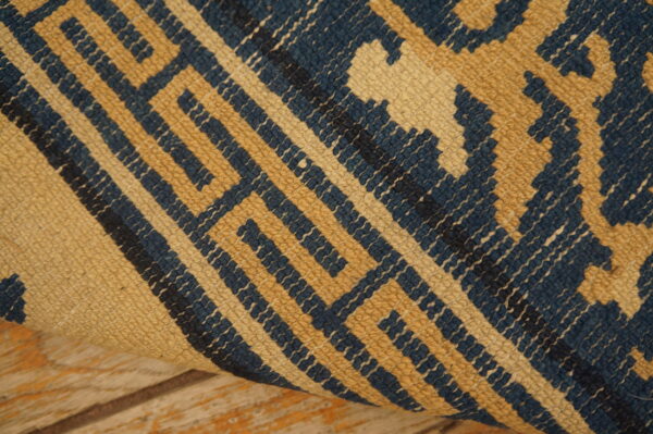 A closeup shows a rolled rug fragment featuring a geometric blue and tan repeating pattern over distressed wood floorboards.