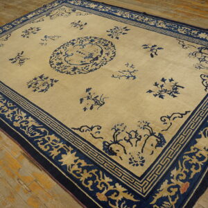 Tan and dark blue rug featuring a sparse floral design and elaborate borders rests on distressed wood flooring.