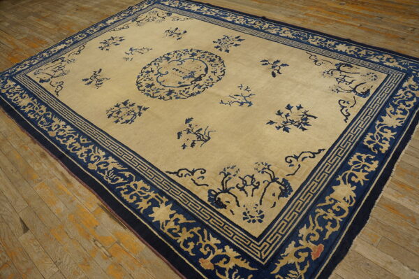 Tan and dark blue rug featuring a sparse floral design and elaborate borders rests on distressed wood flooring.