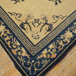Low-pile beige rug with dark blue floral and key pattern borders resting on distressed wooden flooring.