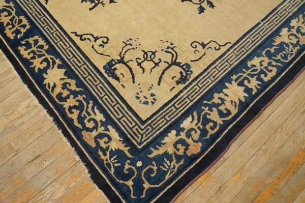 Low-pile beige rug with dark blue floral and key pattern borders resting on distressed wooden flooring.