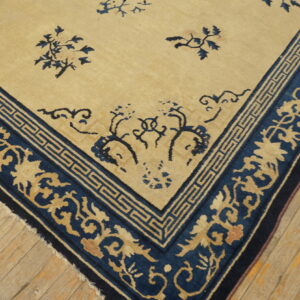 Tan and dark blue rug featuring a scrolling floral and Greek key border on wooden floorboards.
