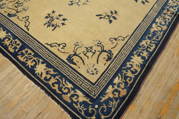 Tan and dark blue rug featuring a scrolling floral and greek key border on wooden floorboards.