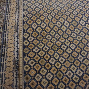 Navy and beige rug with geometric motifs and patterned borders laid over worn golden wooden planks.