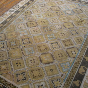 Yellow-gold rug featuring a checkered pattern, blue accents, and dark borders with Greek key designs on wood flooring.