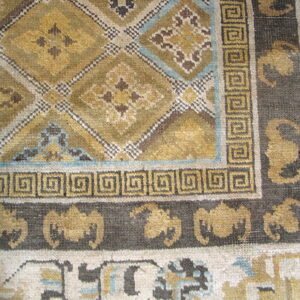 A close-up of a rug corner with gold and olive geometric patterns bordered by dark meander and repeating motifs.