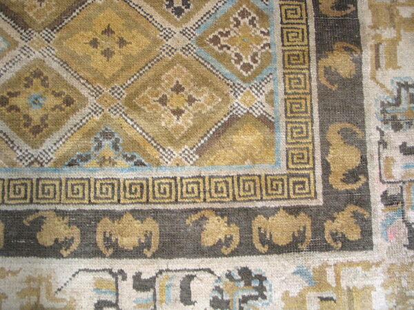 A close-up of a rug corner with gold and olive geometric patterns bordered by dark meander and repeating motifs.