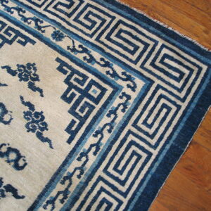 A cream and blue rug with a dense Greek key border pattern rests on warm brown hardwood flooring.