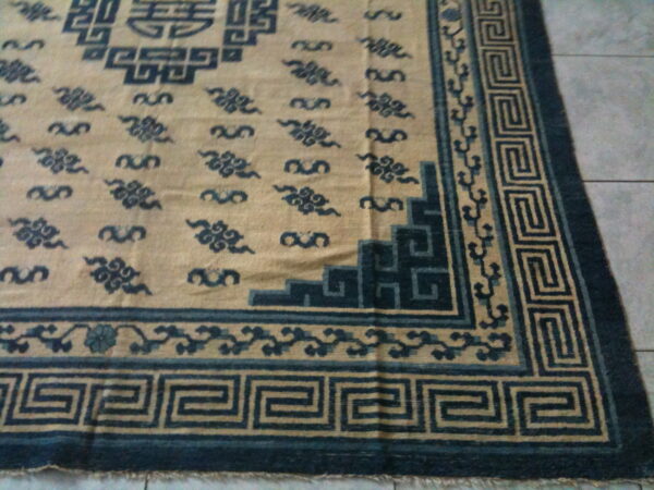 Tan rug featuring dark blue meander borders and scrolling cloud motifs on a gray tile background.