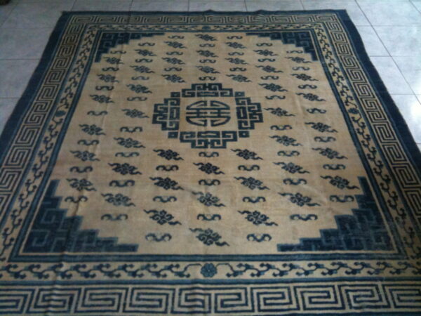 Tan and dark blue rug with a central medallion, scattered cloud motifs, and a greek key border on tile.