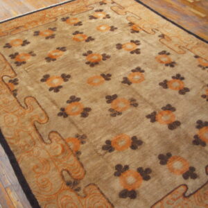 Tan field rug with repeating orange and brown stylized floral motifs and large curvilinear borders on wood flooring.