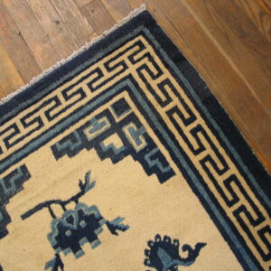 Cream rug with dark blue floral elements and a meander border rests on worn wood flooring.