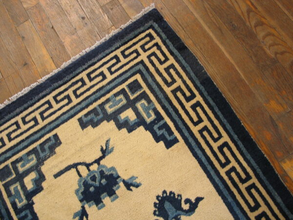 Cream rug with dark blue floral elements and a meander border rests on worn wood flooring.