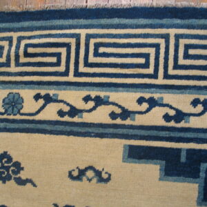 Close-up of a rug border featuring a dark blue key pattern and scrolling motifs on a cream field.