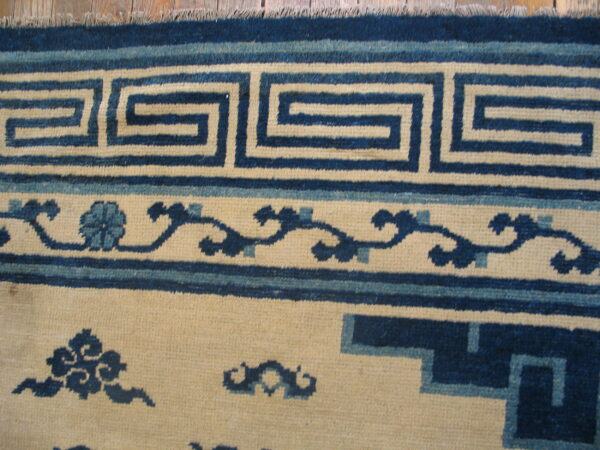 Close-up of a rug border featuring a dark blue key pattern and scrolling motifs on a cream field.