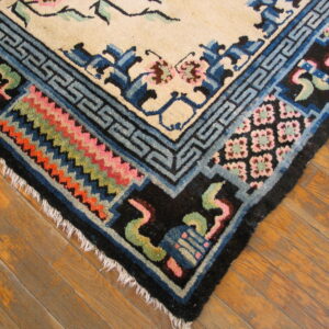 Cream rug corner with blue geometric borders, floral motifs, colorful zigzag patterns, and fringe on wood planks.