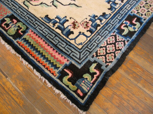 Cream rug corner with blue geometric borders, floral motifs, colorful zigzag patterns, and fringe on wood planks.