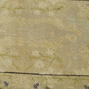 A beige patterned rug with dark motifs rests on concrete next to a person's foot in a sandal.