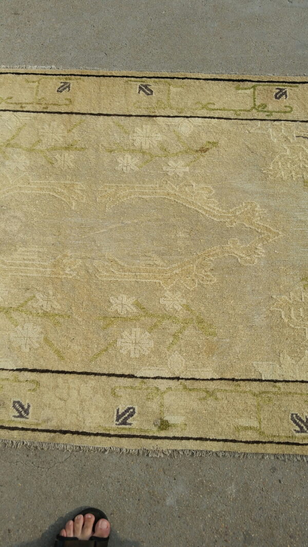 A beige patterned rug with dark motifs rests on concrete next to a person's foot in a sandal.