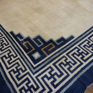 Light beige rug featuring a wide navy blue and cream fretwork border rests on distressed wood flooring.
