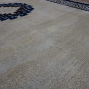 Low-pile neutral rug with dark blue and tan interlocking key patterns and a dark wood floor edge.