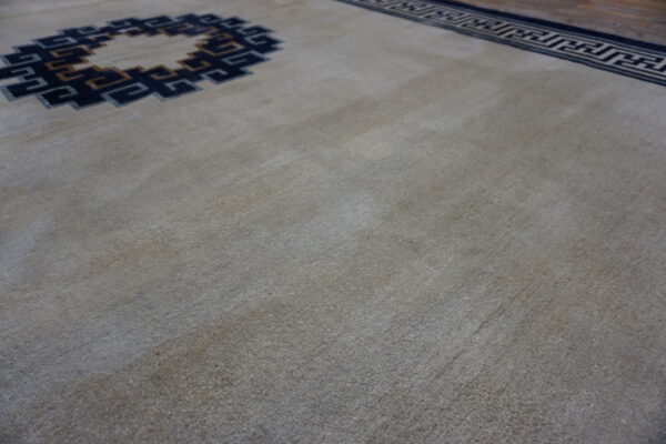 Low-pile neutral rug with dark blue and tan interlocking key patterns and a dark wood floor edge.