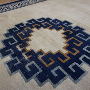 Low pile beige rug with a concentric navy, tan, and gray geometric stepped central medallion.