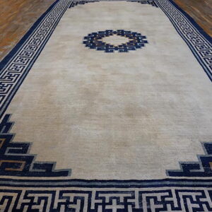 Beige rug with a navy geometric fretwork border and central design on hardwood flooring.