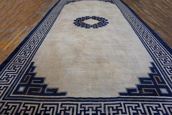 Beige rug with a navy geometric fretwork border and central design on hardwood flooring.