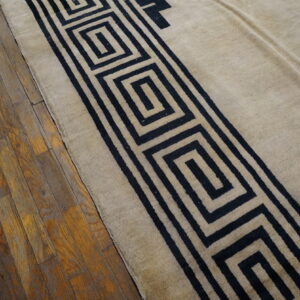 Beige rug with a black geometric Greek key pattern border rests on worn wood plank flooring.