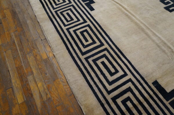 Beige rug with a black geometric greek key pattern border rests on worn wood plank flooring.