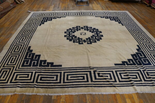 Large beige rug featuring a prominent dark geometric fretwork medallion and greek key border on hardwood flooring.