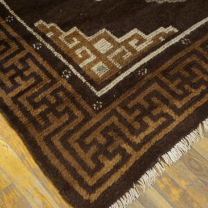 This brown and tan rug displays a thick pile, geometric border, and fringe over worn wood flooring.