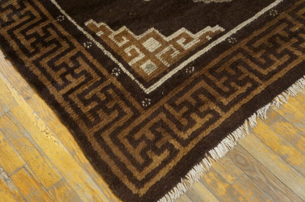 This brown and tan rug displays a thick pile, geometric border, and fringe over worn wood flooring.