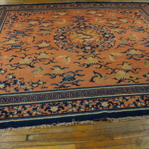 Orange field rug with complex navy and cream floral patterns and an ornate Greek key border on wood floor.