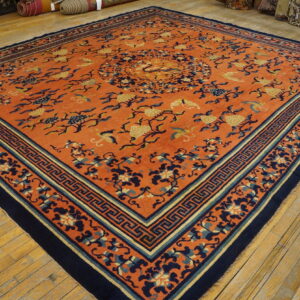 A vibrant orange rug featuring a navy blue and gold floral pattern and key border on wooden planks.