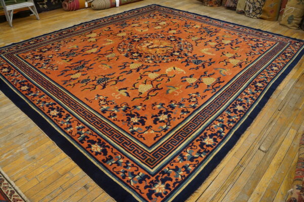 A vibrant orange rug featuring a navy blue and gold floral pattern and key border on wooden planks.