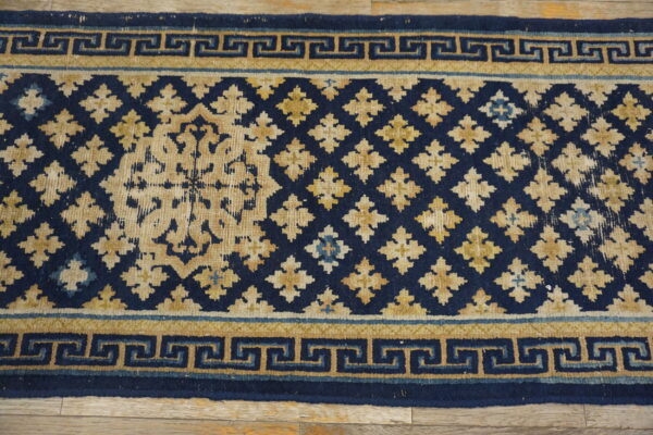 Low-pile navy and tan rug showing a geometric lattice field and greek key border on wood flooring.