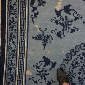 Dusty blue rug with navy floral designs and geometric borders rests on wood flooring, showing a black shoe.