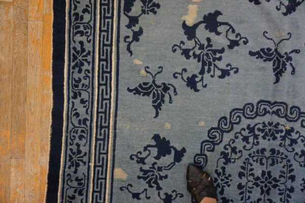 Dusty blue rug with navy floral designs and geometric borders rests on wood flooring, showing a black shoe.