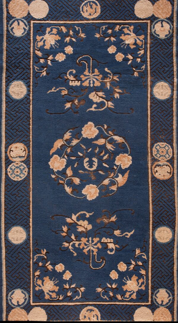 Detail of a blue rug with tan floral designs and a border featuring brown fretwork and circular medallions.