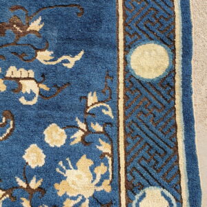 Close-up of a blue rug featuring beige and brown floral patterns and a geometric border resting on concrete.