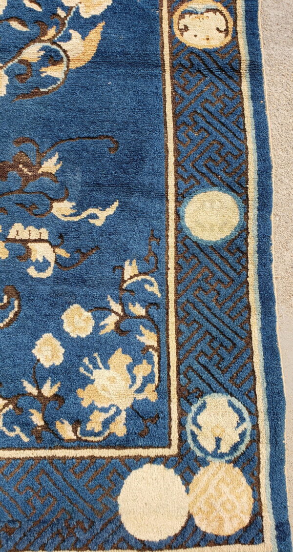 Close-up of a blue rug featuring beige and brown floral patterns and a geometric border resting on concrete.