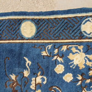 Blue, cream, and brown high-pile rug featuring florals and a geometric key border on concrete.