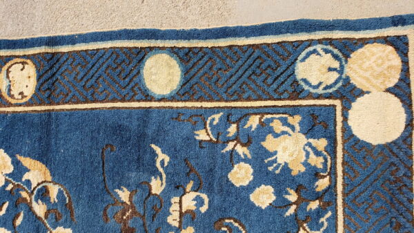 Blue, cream, and brown high-pile rug featuring florals and a geometric key border on concrete.