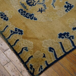 Corner detail of a gold rug featuring navy blue and beige abstract cloud patterns on worn hardwood.