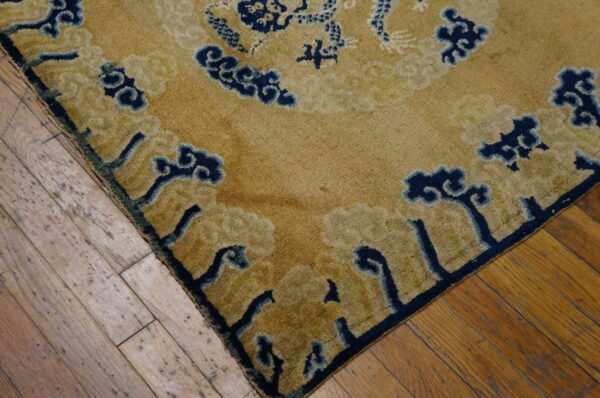 Corner detail of a gold rug featuring navy blue and beige abstract cloud patterns on worn hardwood.