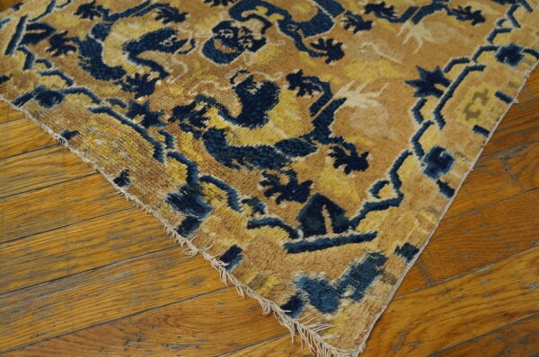 Shaggy blue and yellow patterned rug corner shows detailed motifs and short frayed fringe on wood flooring.