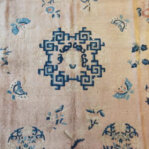 A muted pink rug features a navy geometric key pattern medallion surrounded by scattered blue floral and butterfly motifs.